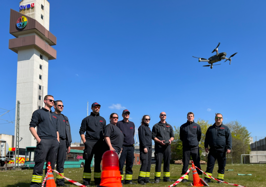 Feuerwehrteam in uniform, steht in einer Reihe, während eine Drohne darüber fliegt.