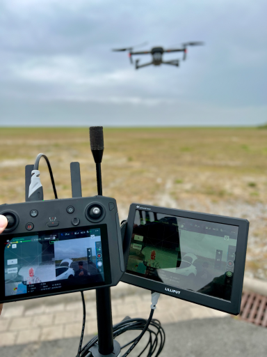 Drohne fliegt über offenes Feld, mit Fernsteuerung und Monitor im Vordergrund.